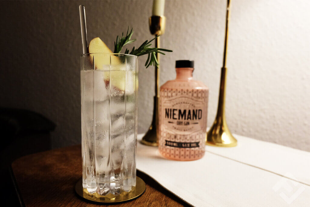 A gin and tonic with an apple slice and a sprig of rosemary and a bottle of Niemand Gin on a table.