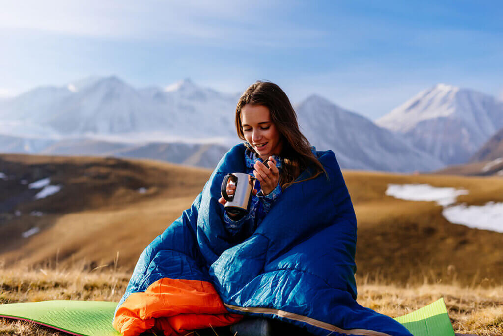 Woman sitting in a mountain landscape wrapped in a sleeping bag and drinking a hot drink